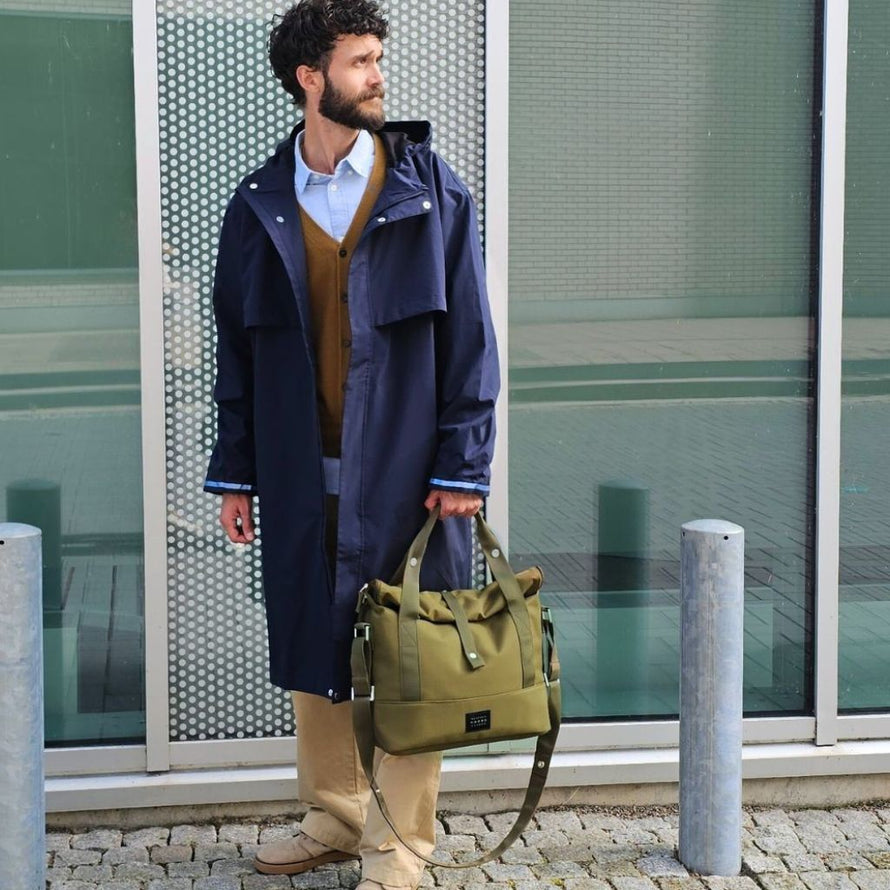Homme en tenue urbaine portant la veste Imbris marine ouverte, avec un sac vert à la main, prêt pour un déplacement en ville.